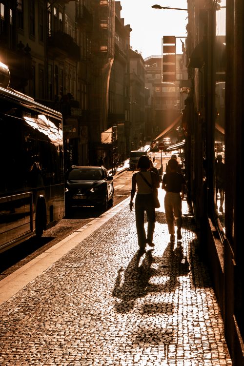 Free Two people walking along a sunlit cobblestone street in Porto, Portugal at sunset, casting long shadows. Stock Photo