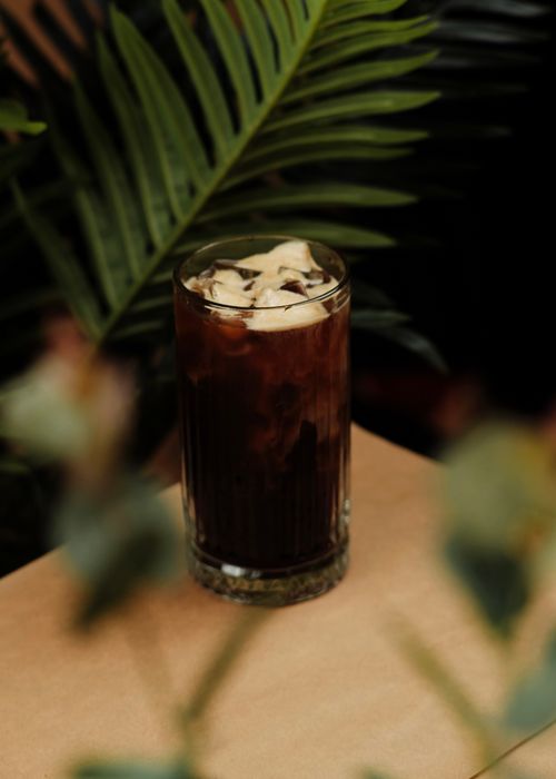 Free A glass of creamy iced coffee on a table with lush green tropical leaves in the background. Stock Photo