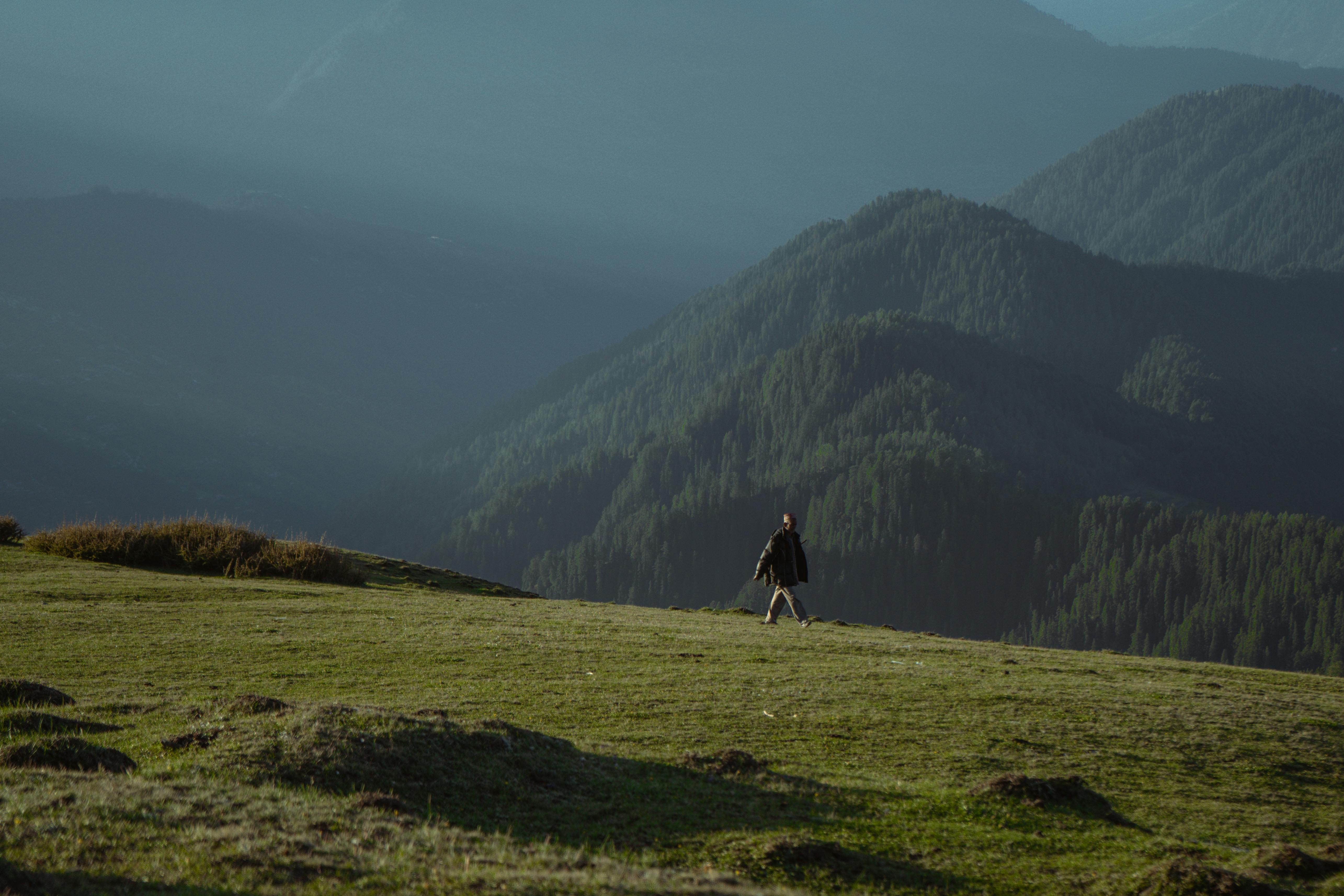 A lone hiker walks through lush greenery with the stunning Shoja hills in the background.