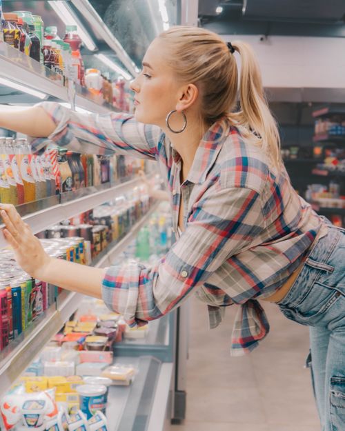 Free Young woman reaches for a drink on a supermarket shelf. Casual and stylish shopping scene. Stock Photo