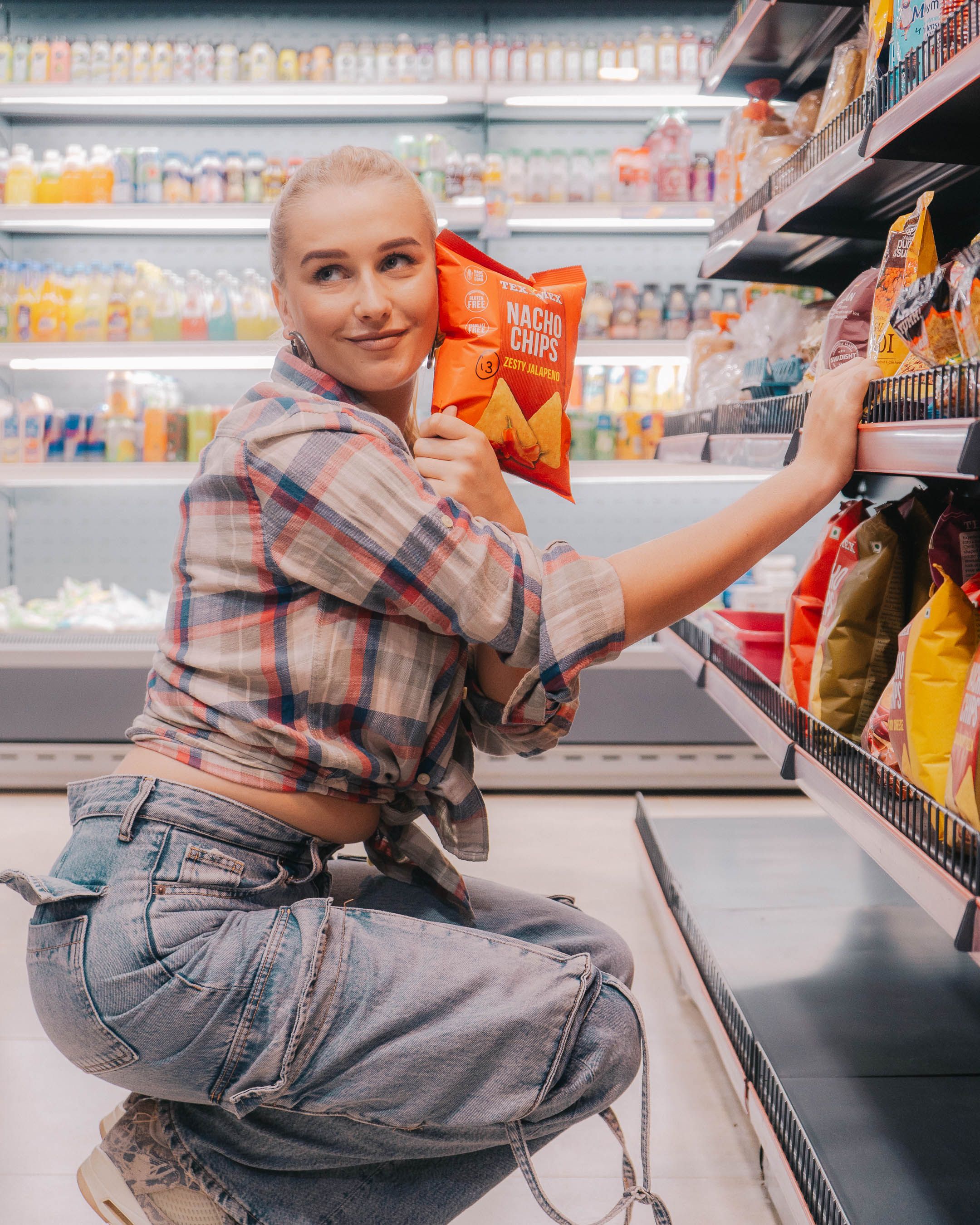 Smiling woman in plaid shirt holding nacho chips bag in a supermarket aisle.