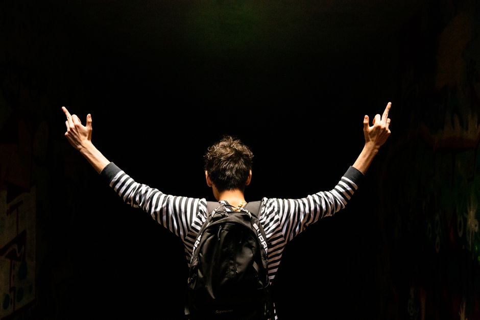 Back view of a man in a striped shirt and backpack giving a hand gesture in a dark tunnel.