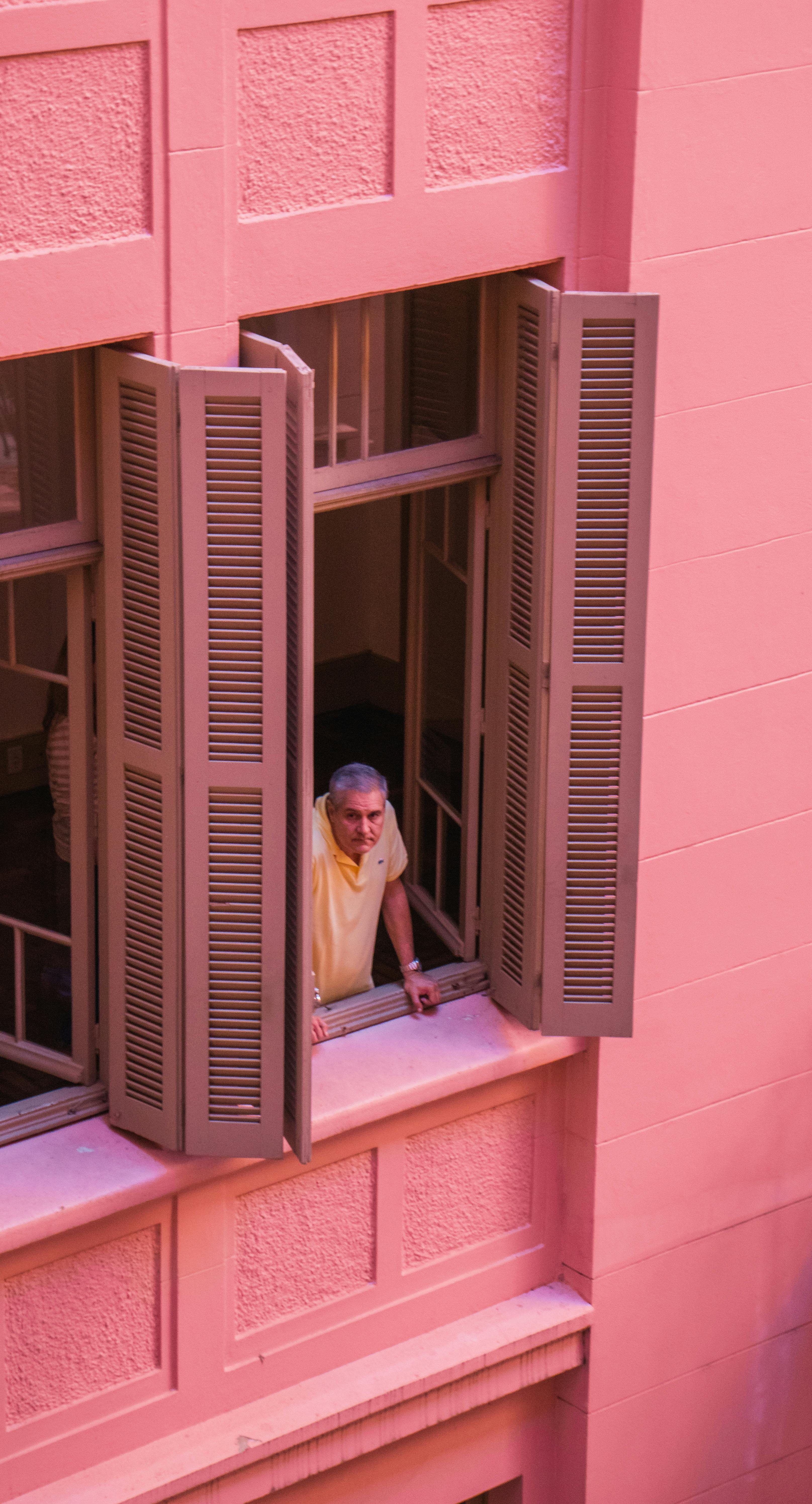 High angle of senior man looking out of large window of pink house in city while spending time at home