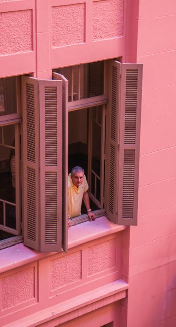 High angle of senior man looking out of large window of pink house in city while spending time at home