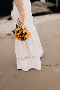 Woman in a white dress walking on the beach holding a bright sunflower bouquet.