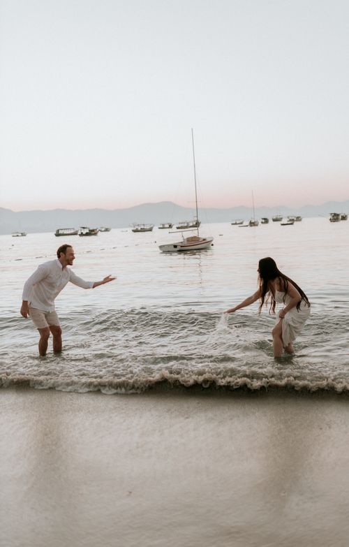 Free A couple playfully splashes in the waves with sailboats in the background during sunset. Stock Photo