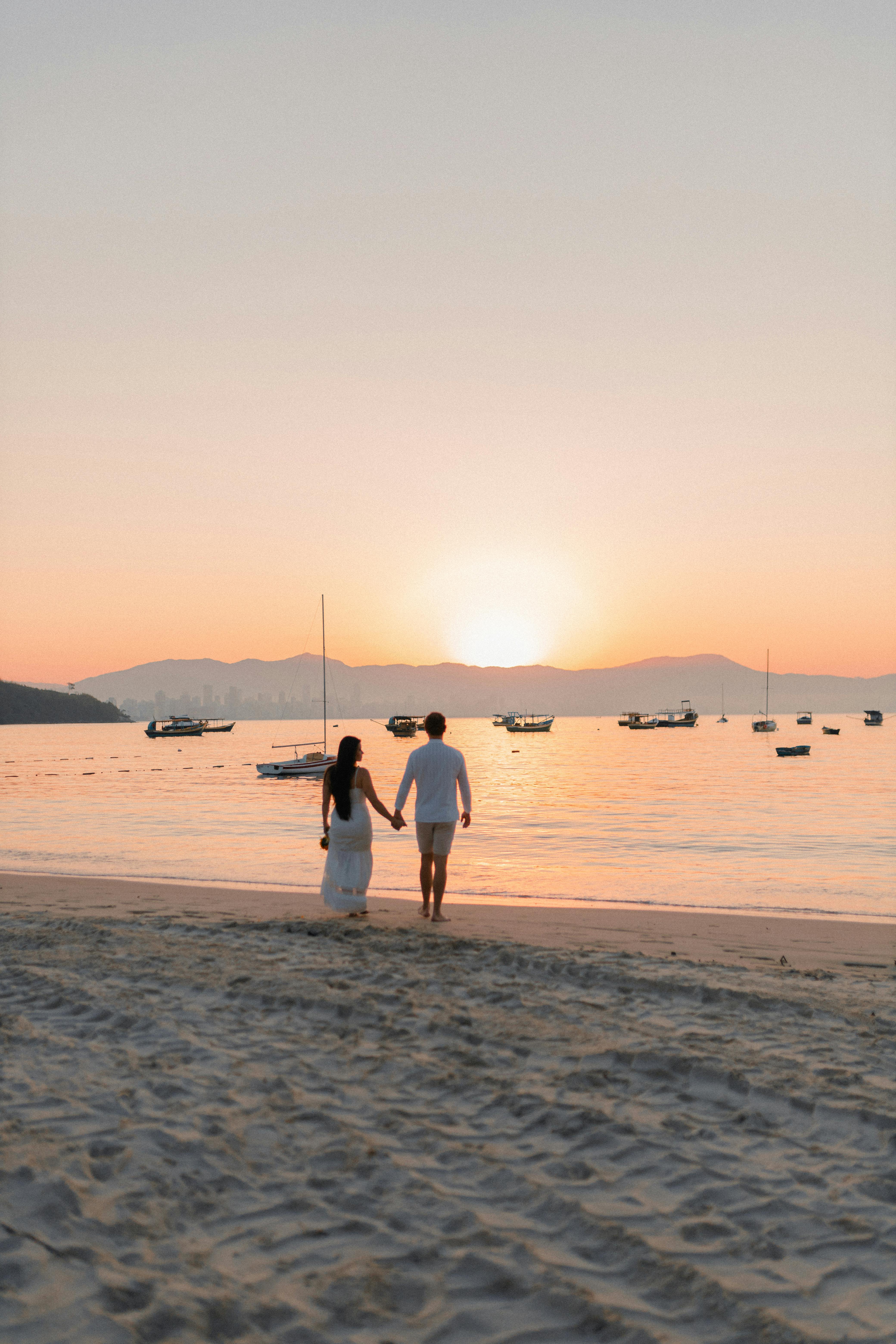 A couple walks hand in hand on a tranquil beach at sunset with boats on the horizon.