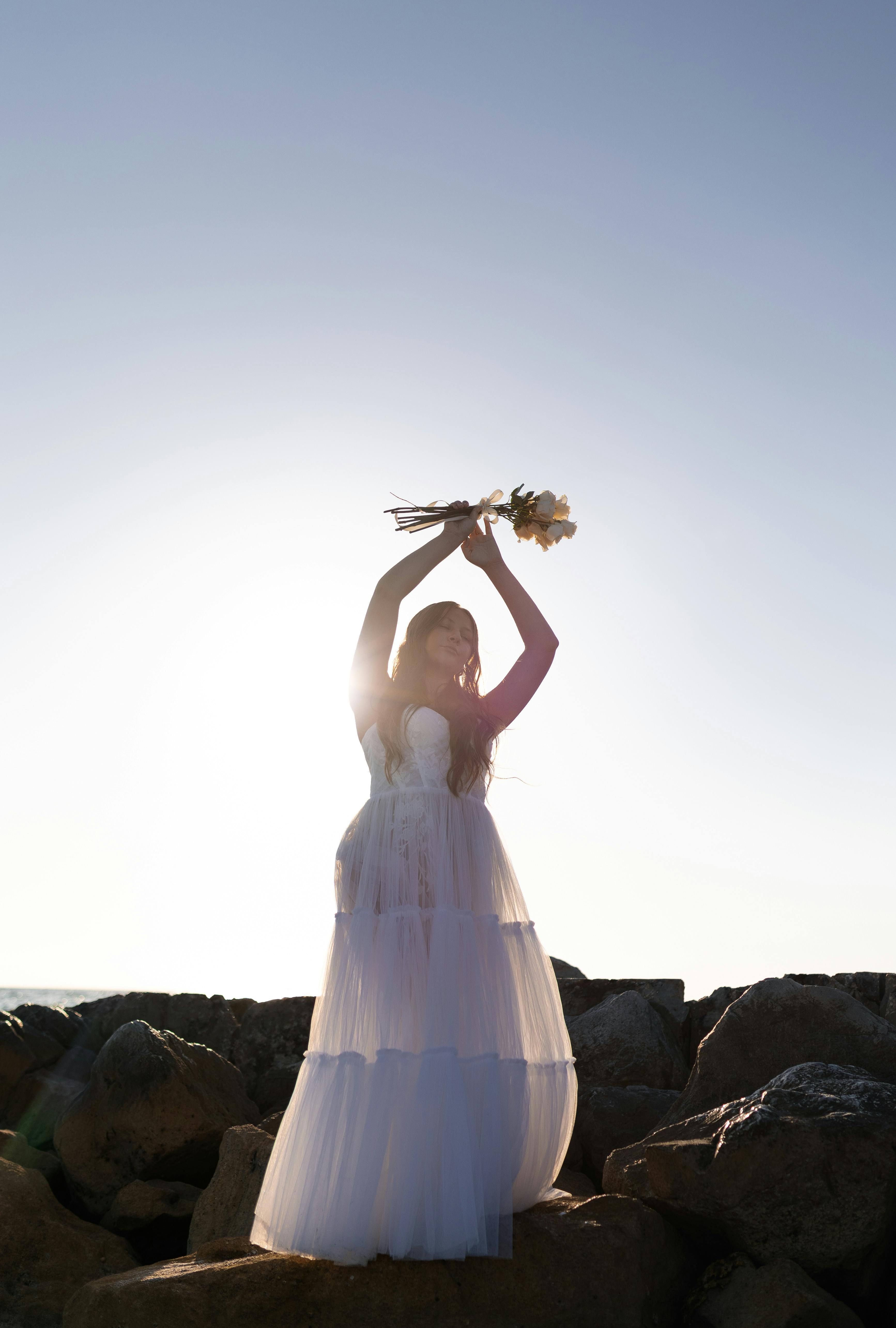A woman in a white dress holding flowers stands on rocks during sunset, creating a serene silhouette.