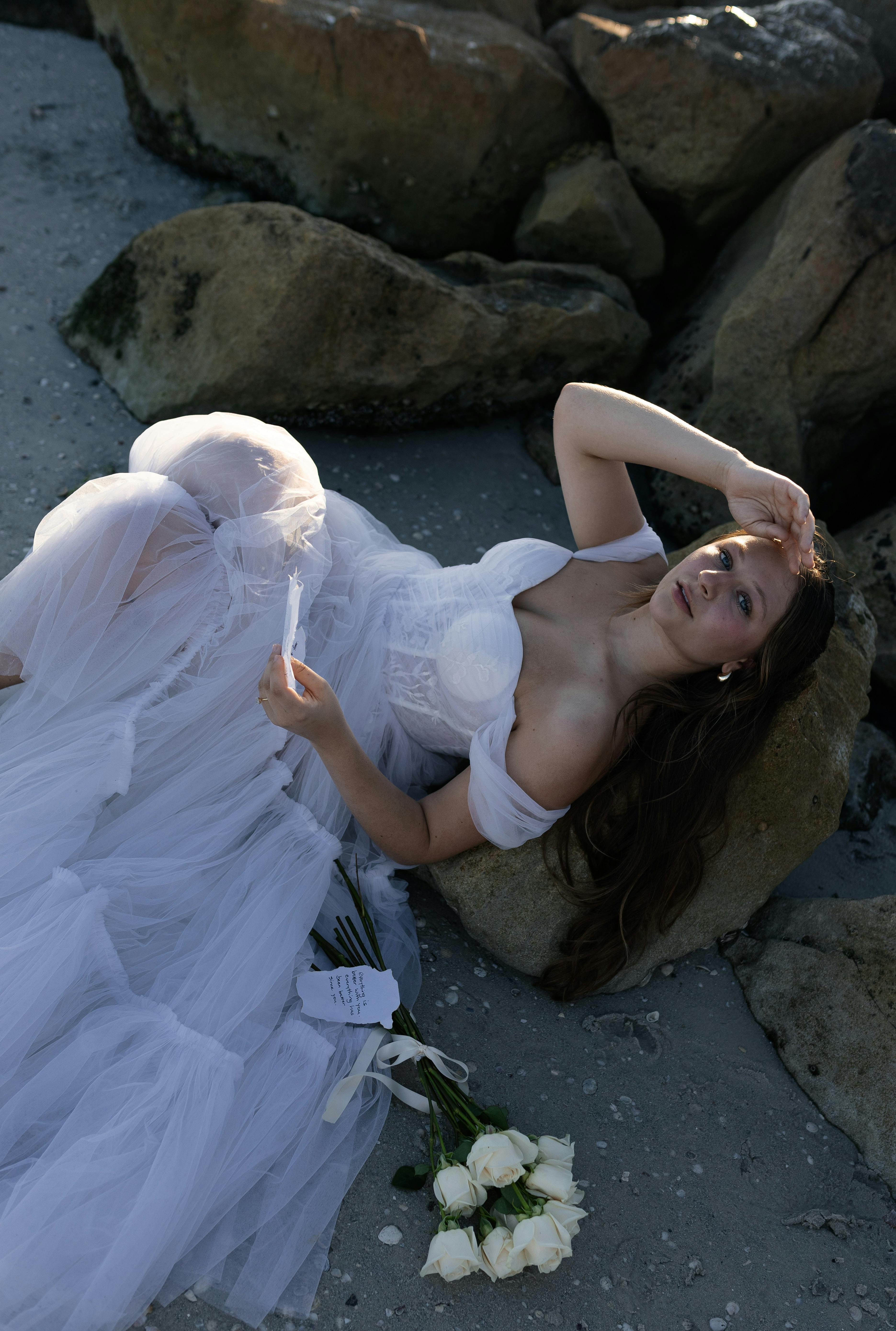 Bride in a white wedding dress relaxing on a rocky beach with a bouquet of roses.