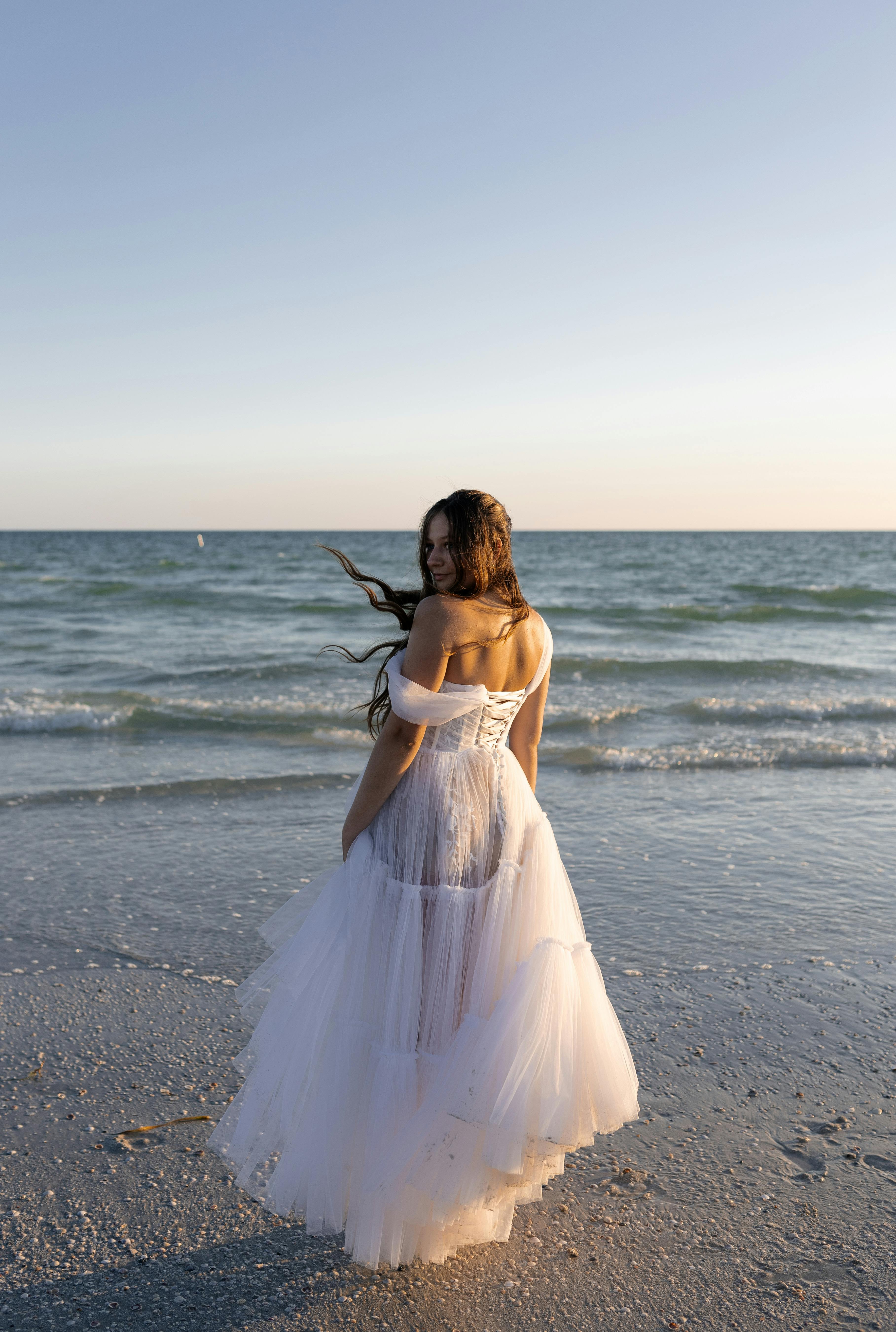 Bride in flowing gown stands on sandy beach at sunset, capturing elegance and romance.