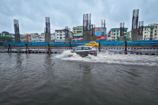 Cars navigate a flooded street in Kolkata, India, under overcast skies.
