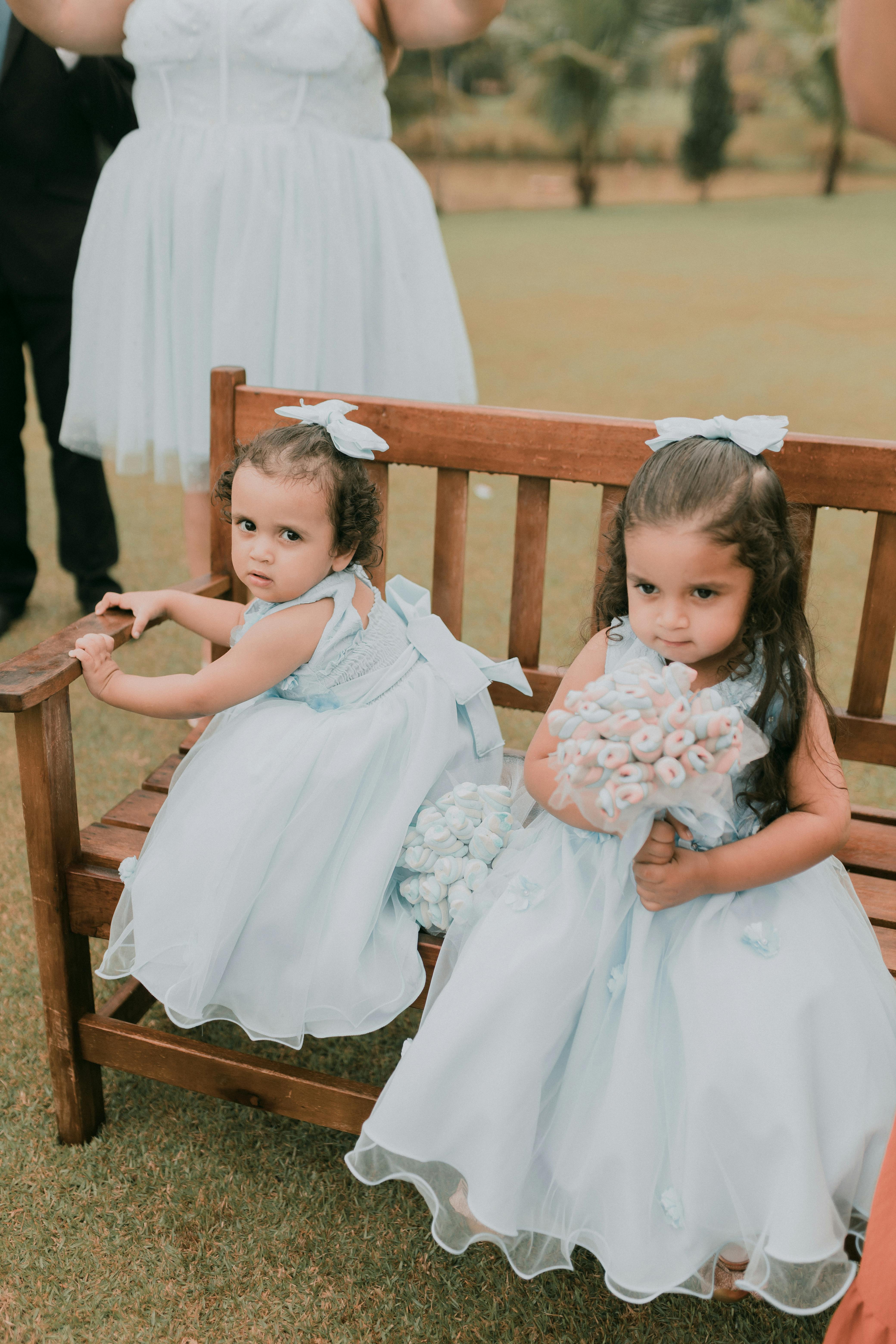 Two young flower girls in blue dresses with bouquets sitting on a wooden bench outdoors at a special occasion.