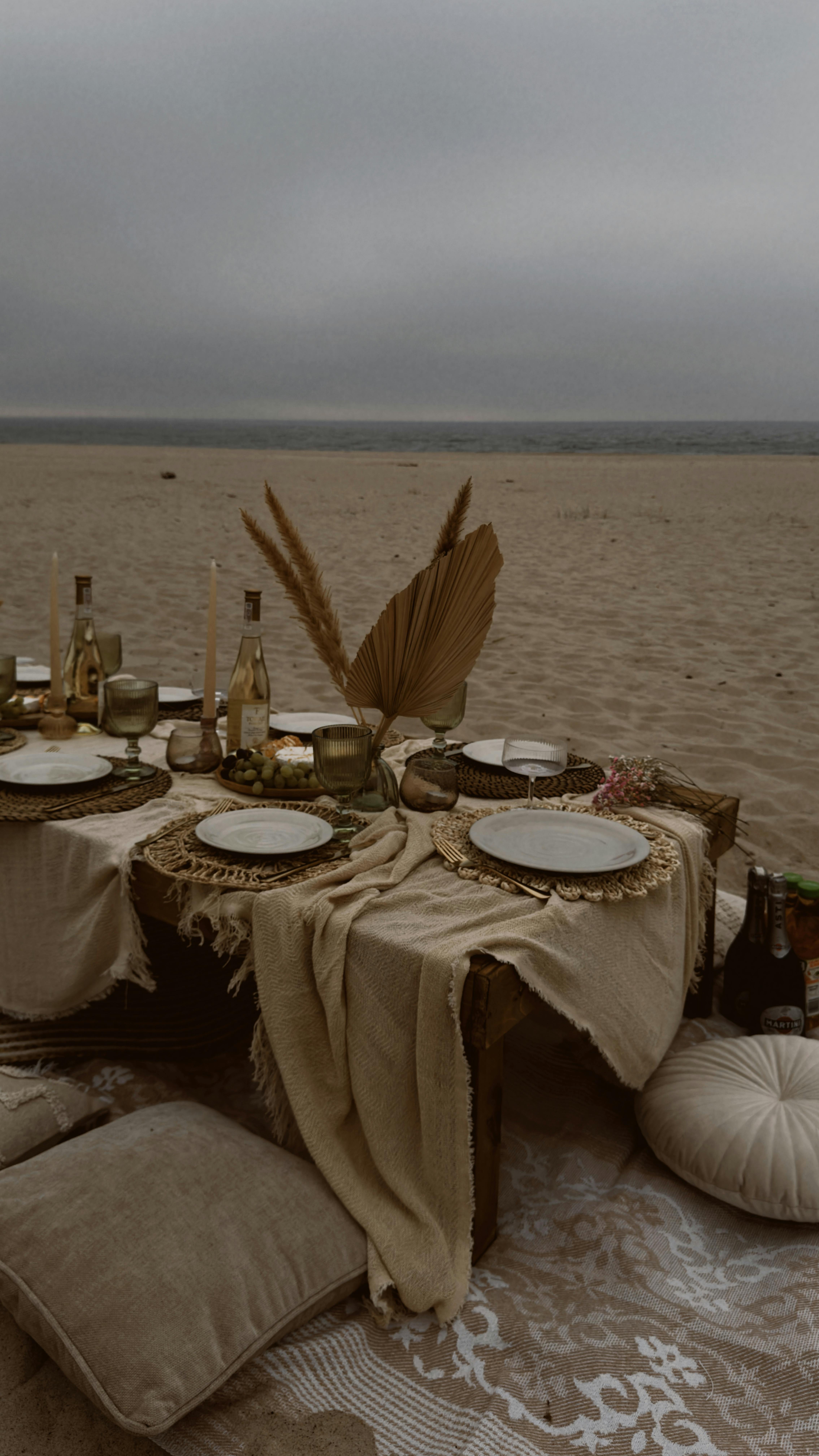 A beautifully set dining table on a beach with elegant tableware and cushions.