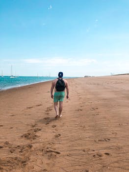 一名男子背着背包，在晴朗的天空下沿着宁静的沙滩散步，享受着夏日的时光