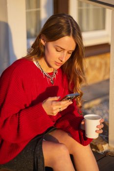 A young woman in a red sweater checks her phone while enjoying coffee in the sunlight.