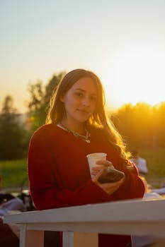 Woman in red sweater holding cup during sunset outdoors.