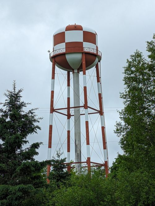 Free A striking red and white water tower stands tall against a cloudy sky, surrounded by lush greenery. Stock Photo