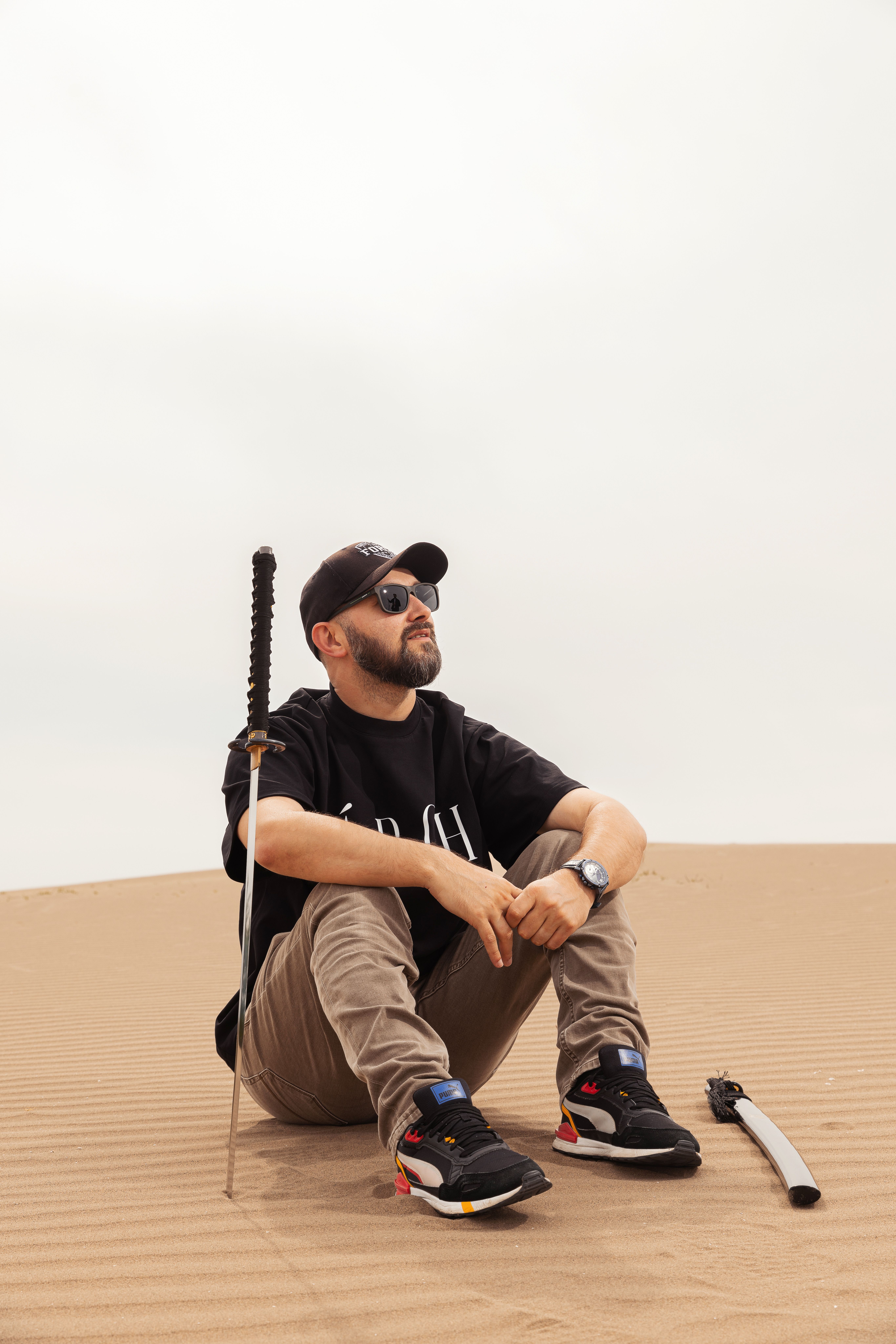 A man in sunglasses and a cap sits on sand in a desert with a samurai sword.