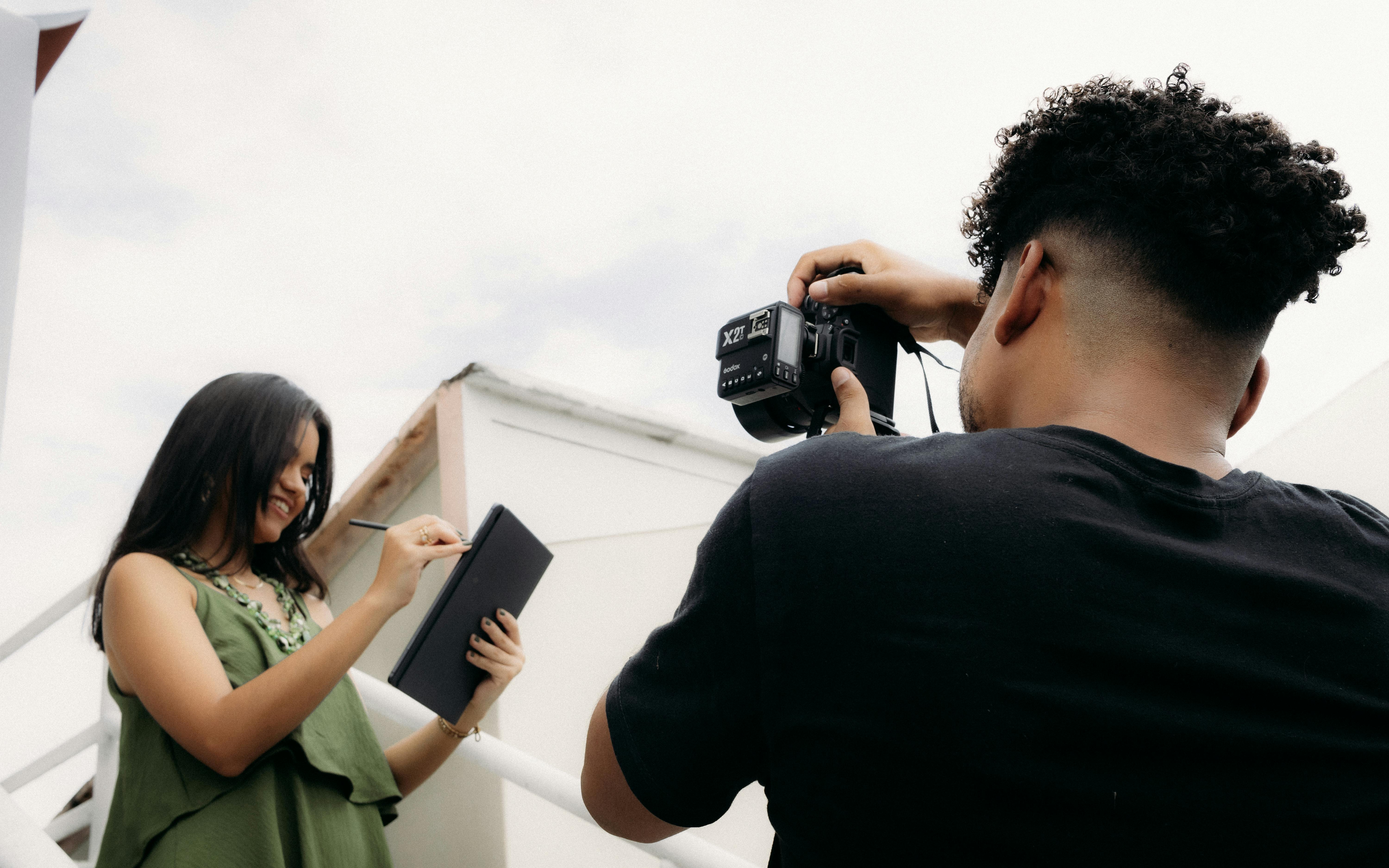 Photographer taking a picture of a smiling young woman holding a notebook outdoors.