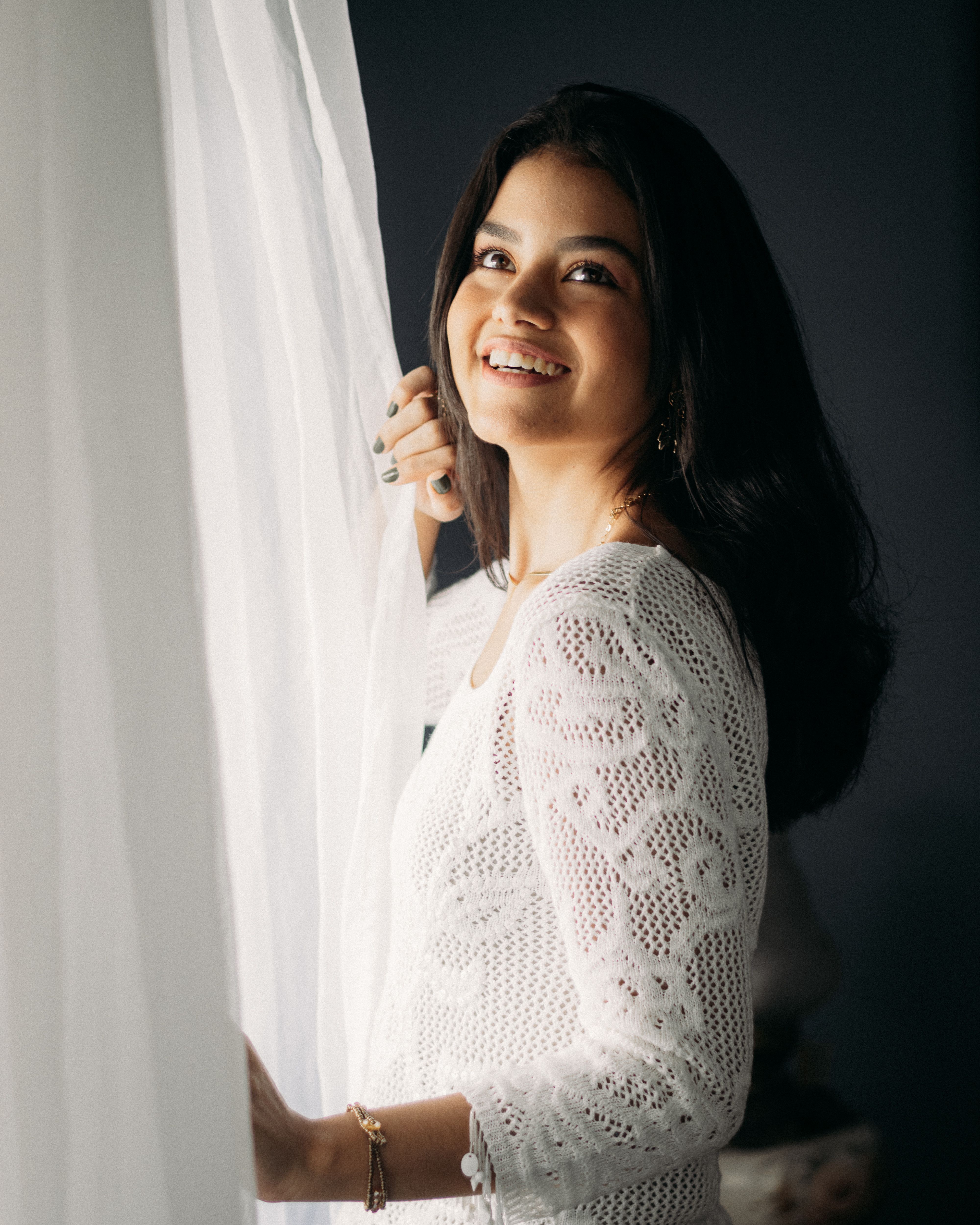 A young woman smiles gracefully by a window in a lace outfit, capturing warmth and elegance indoors.