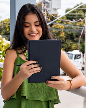 Young woman with black hair and green top smiling while using a tablet outdoors.