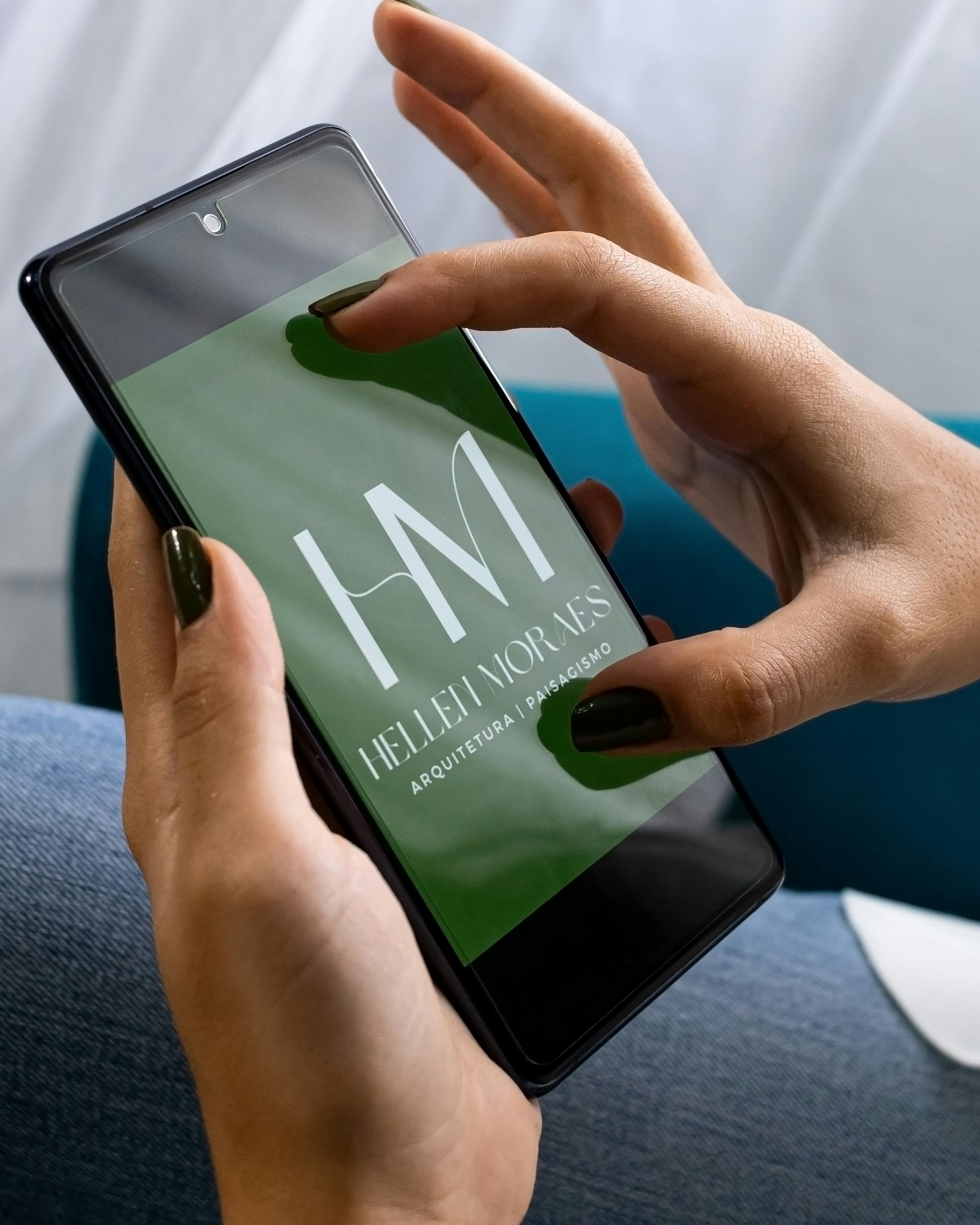 Close-up of hands holding a smartphone with Hellen Moraes logo, showing design and branding concept.