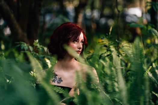 A red-haired woman with tattoos amidst lush green ferns in a forest setting, conveying a sense of natural beauty.