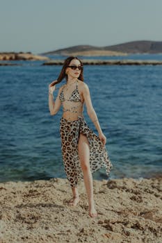 Young woman standing on a rocky beach in a leopard print bikini, enjoying a sunny day.