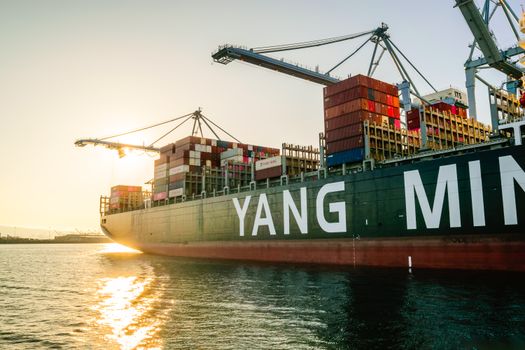 Large cargo ship with stacked containers and cranes at sunset in a harbor.