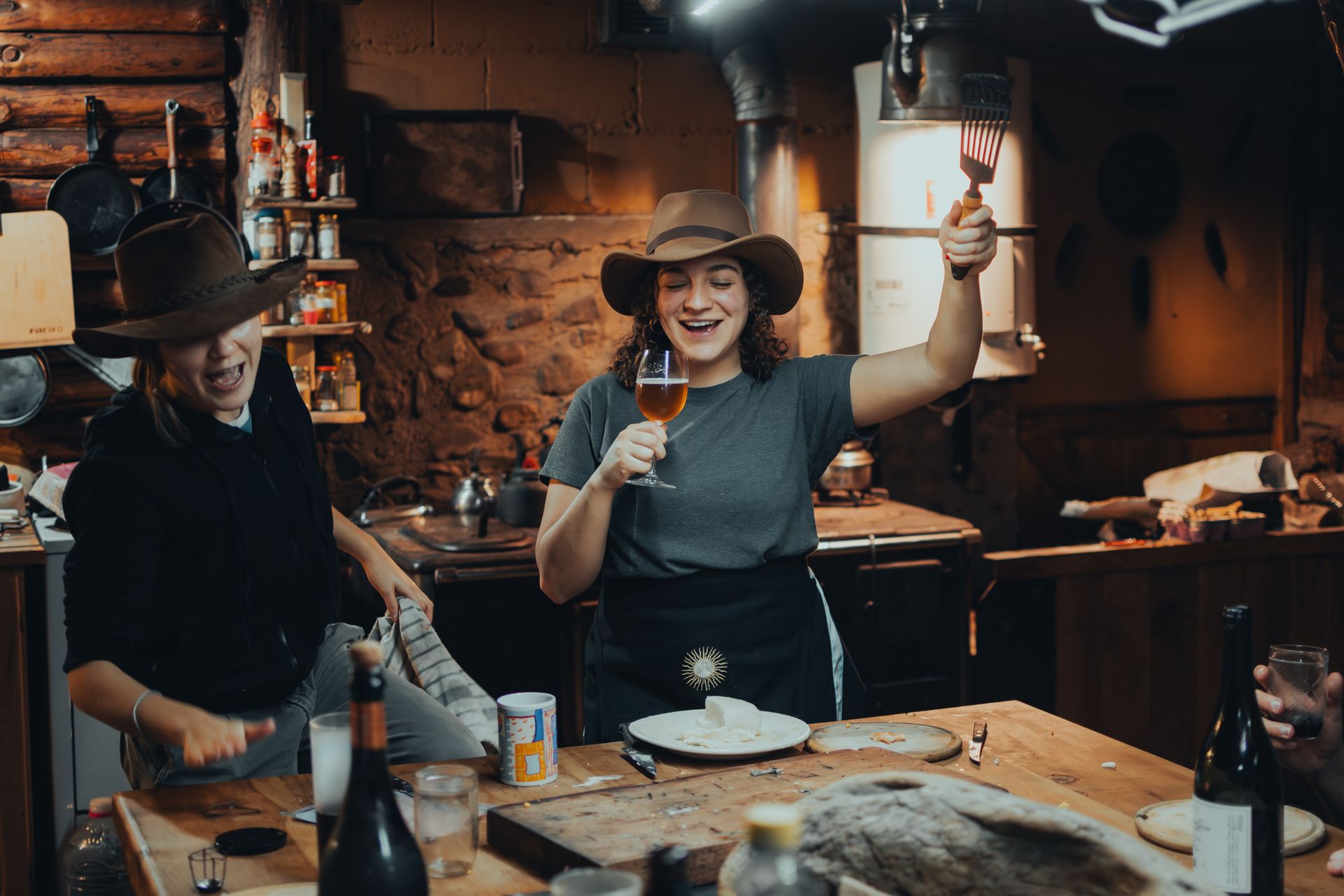 Two women enjoying drinks and cooking in a cozy kitchen setting, filled with laughter.