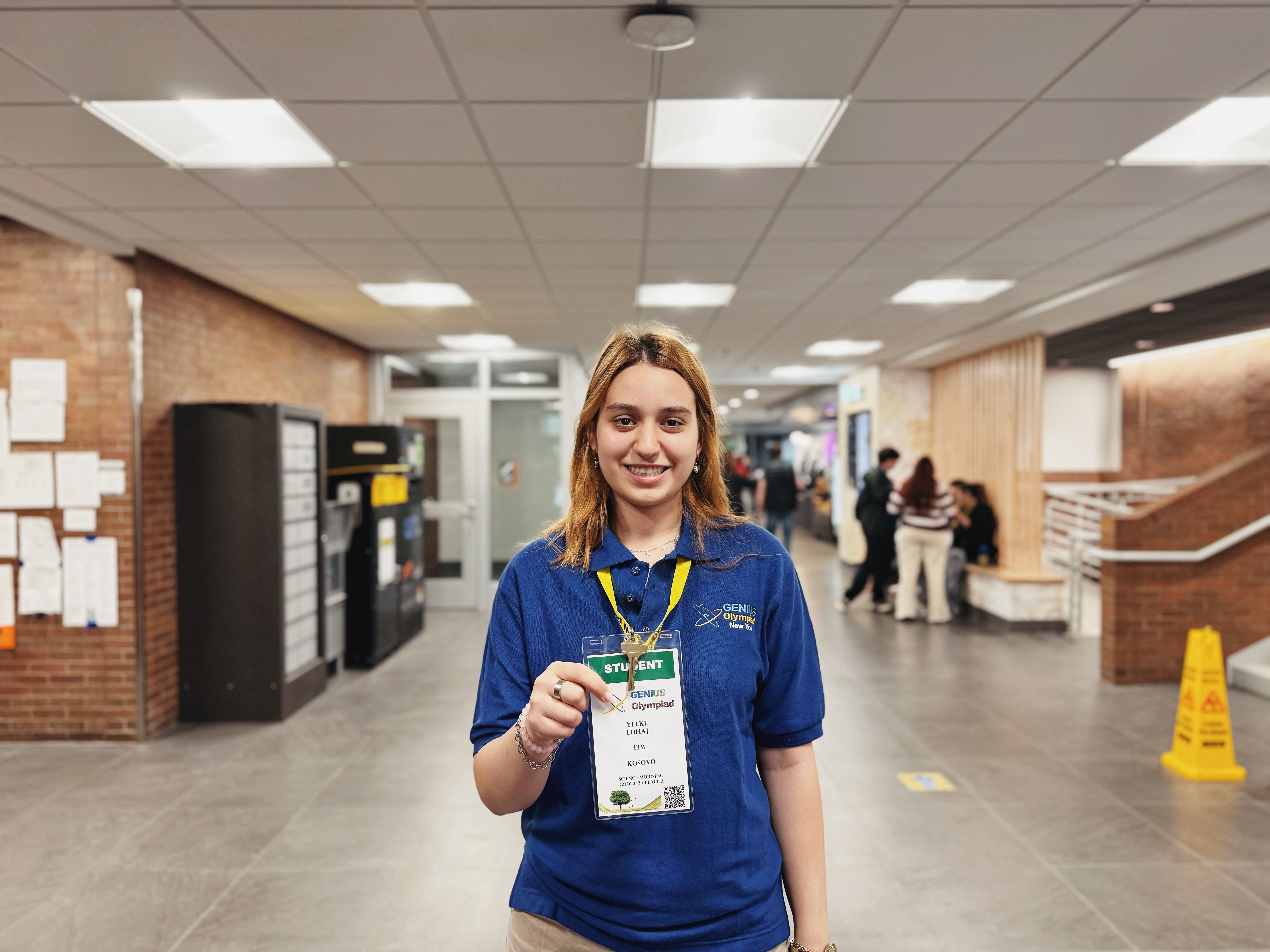 A young student smiling and displaying a badge while standing in a school corridor.