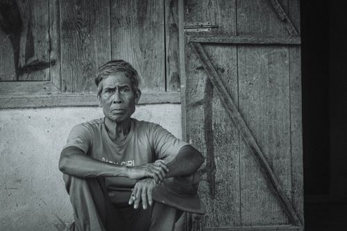 Free Black and white portrait of an elderly man sitting pensively by a rustic door. Stock Photo