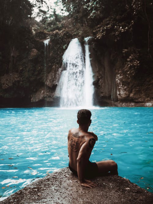 Free Back View of a Topless Man Sitting Across the Waterfalls Stock Photo