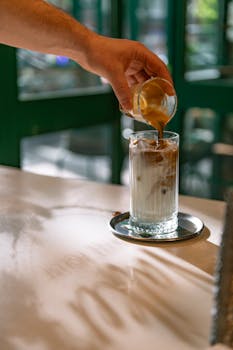 A barista hand pours espresso over iced milk in a glass, creating a layered coffee drink.