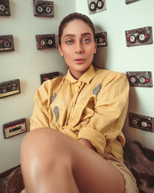 Photo of Woman in Yellow Shirt Leaning on Wall With Cassette Tapes