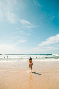 A woman in a pink swimsuit walks towards the ocean on a sunny day, capturing the essence of summer beach vibes.