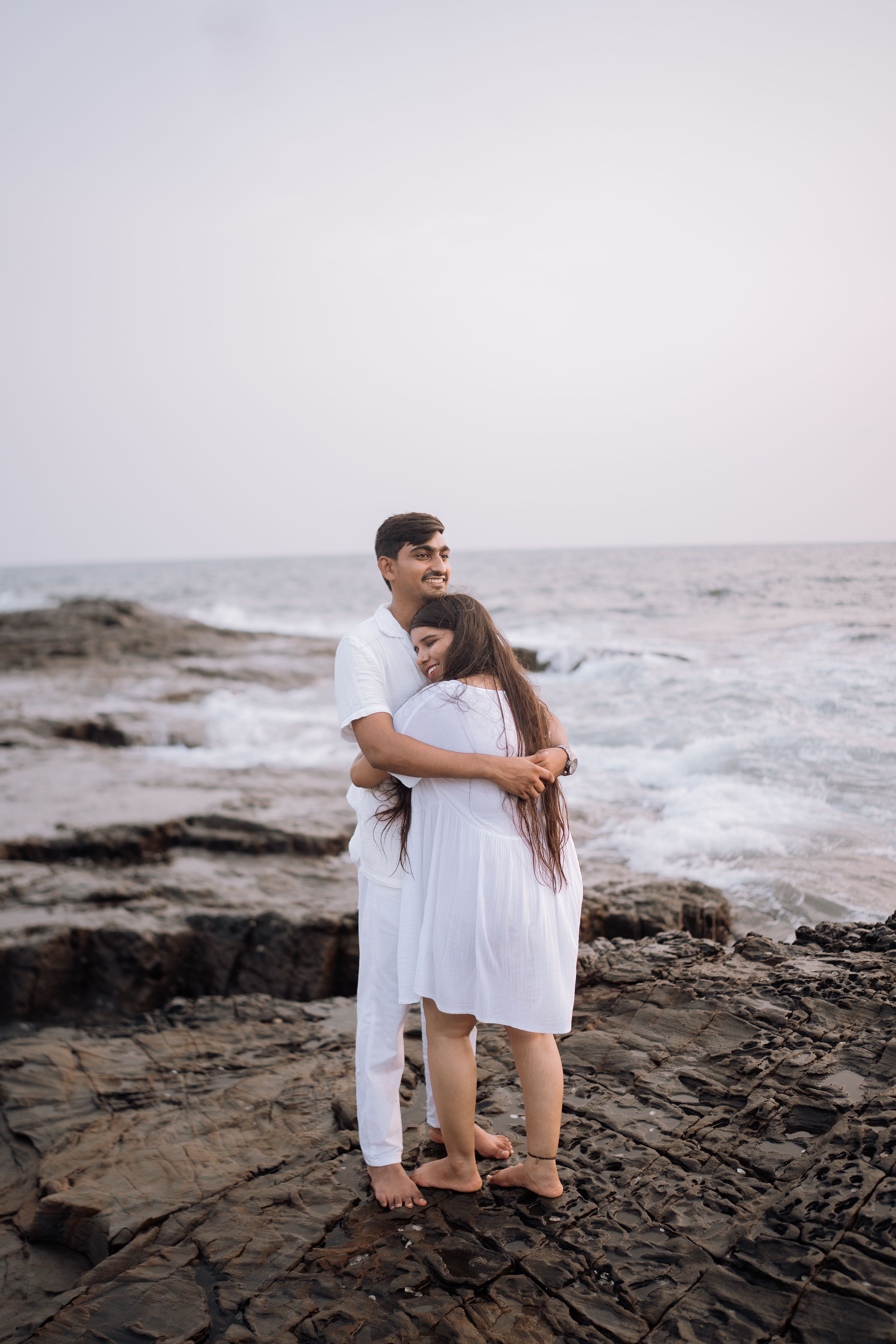 A loving couple in white outfits shares an embrace on the rocky shore of Baga Beach, India.