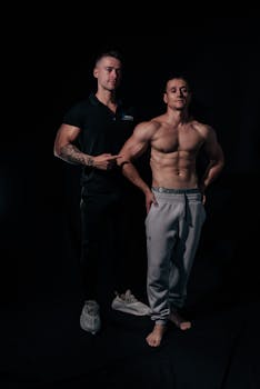Two athletic men, one shirtless, posing in a dimly lit studio setting, showcasing muscular physiques.