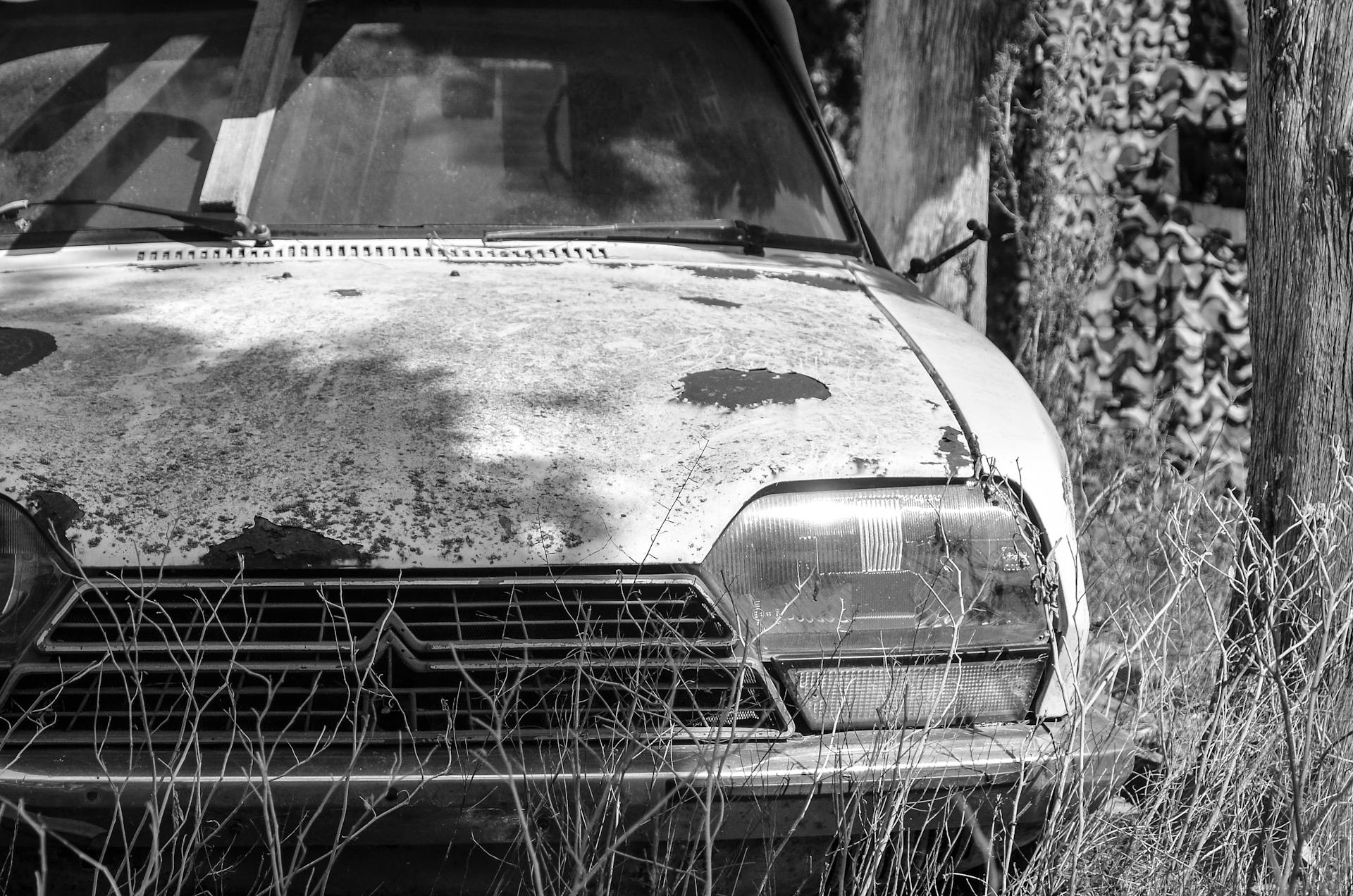 Black and white photo of a decaying, rusty car in an overgrown field.