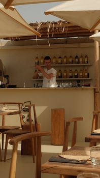 Bartender mixing drinks at an outdoor bar with wood decor under sunny skies.