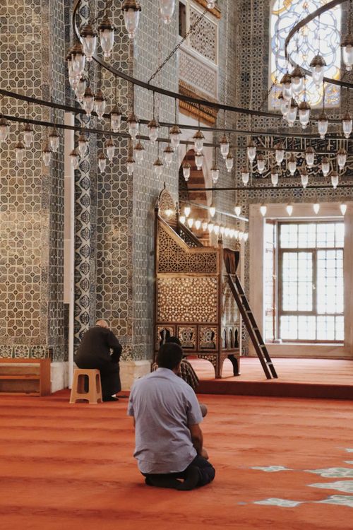 Free Men kneeling and praying inside a richly decorated mosque with intricate tilework and chandeliers. Stock Photo