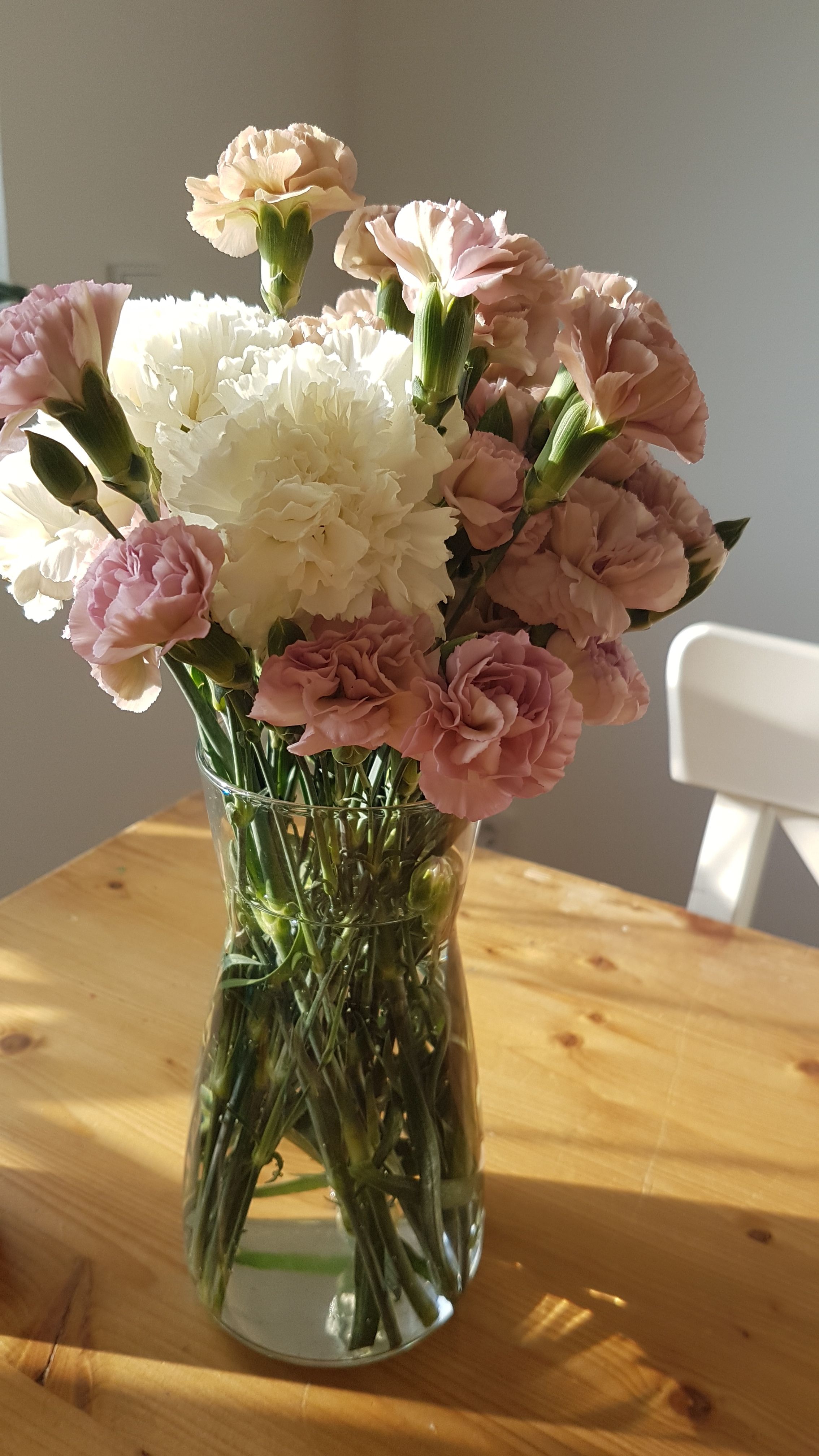 A sunlit vase with pink and white carnations on a wooden table, casting soft shadows.
