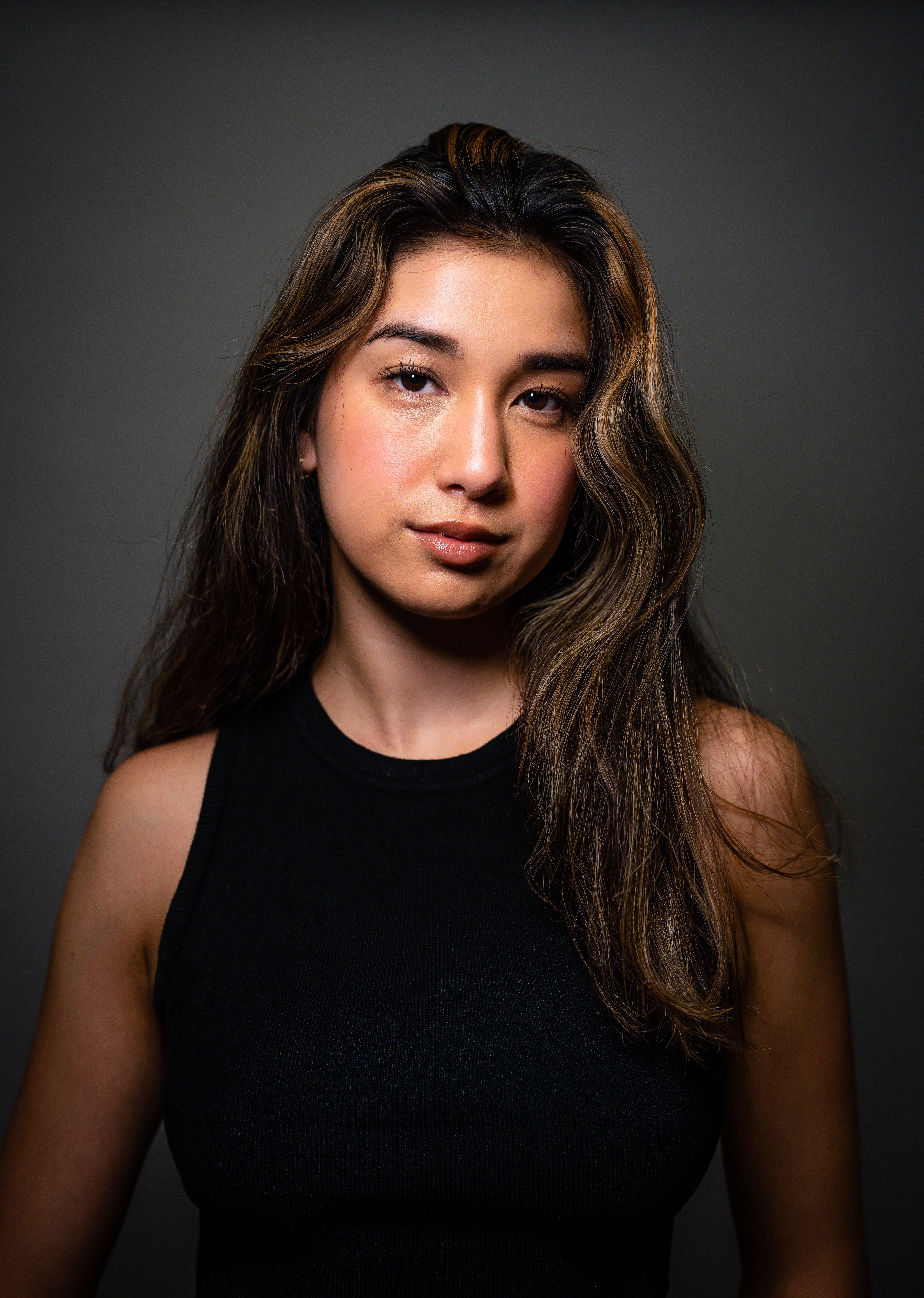 Studio portrait of a young woman with long brunette hair posing confidently against a gray background.
