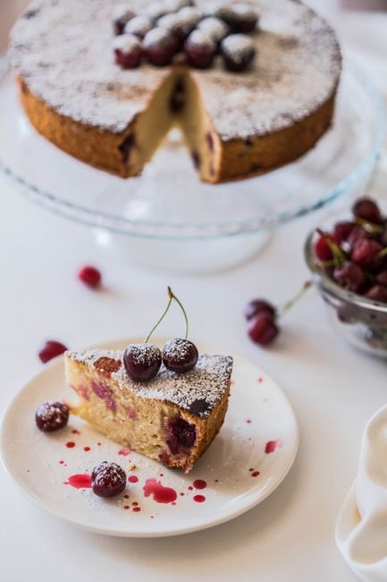 Close-up of a cherry cake slice and whole cake with powdered sugar and cherries on top.