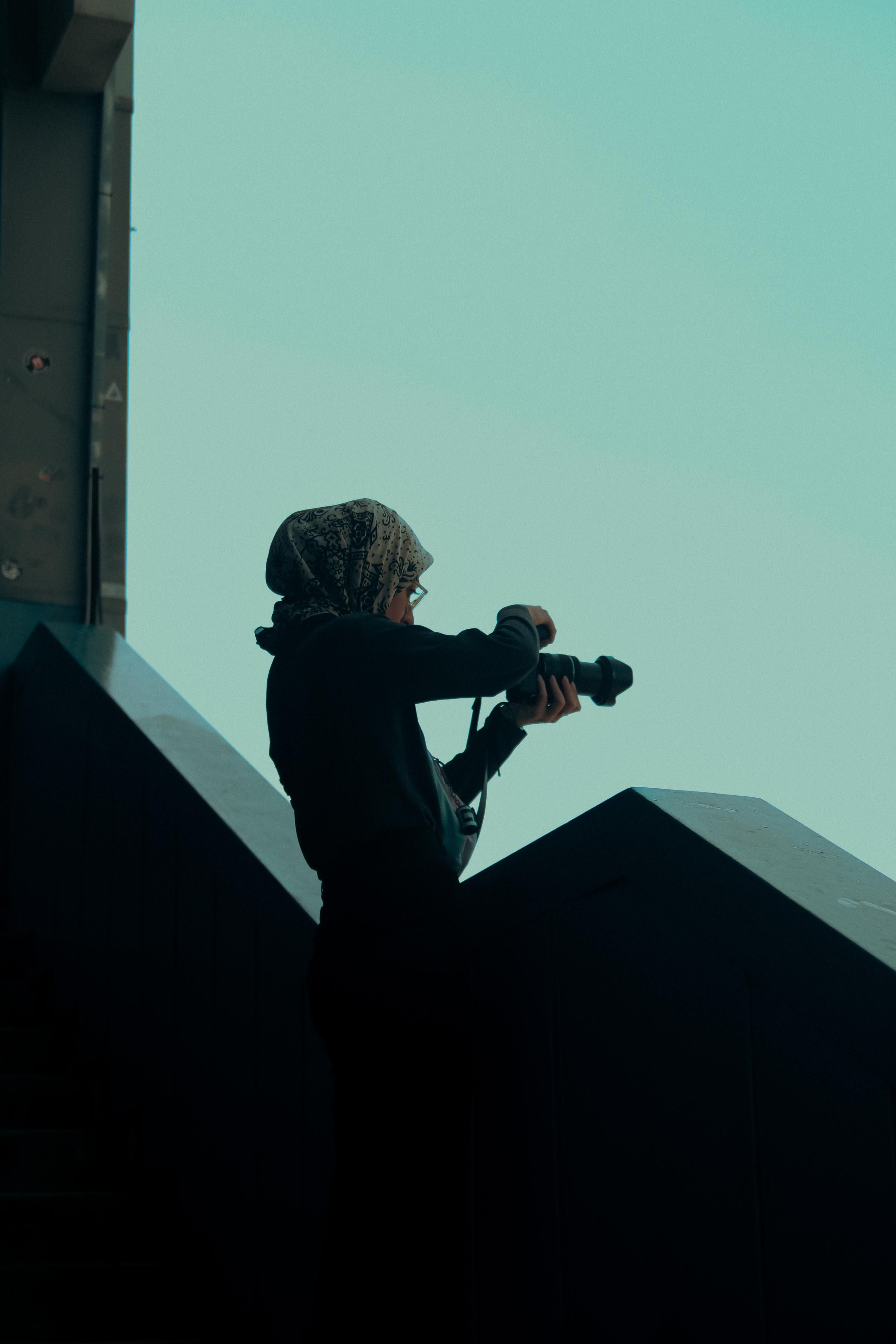 Silhouetted woman with headscarf photographing outdoors in urban setting.