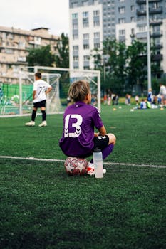 Child sitting on a soccer ball at an urban sports field, wearing sportswear, number 13.