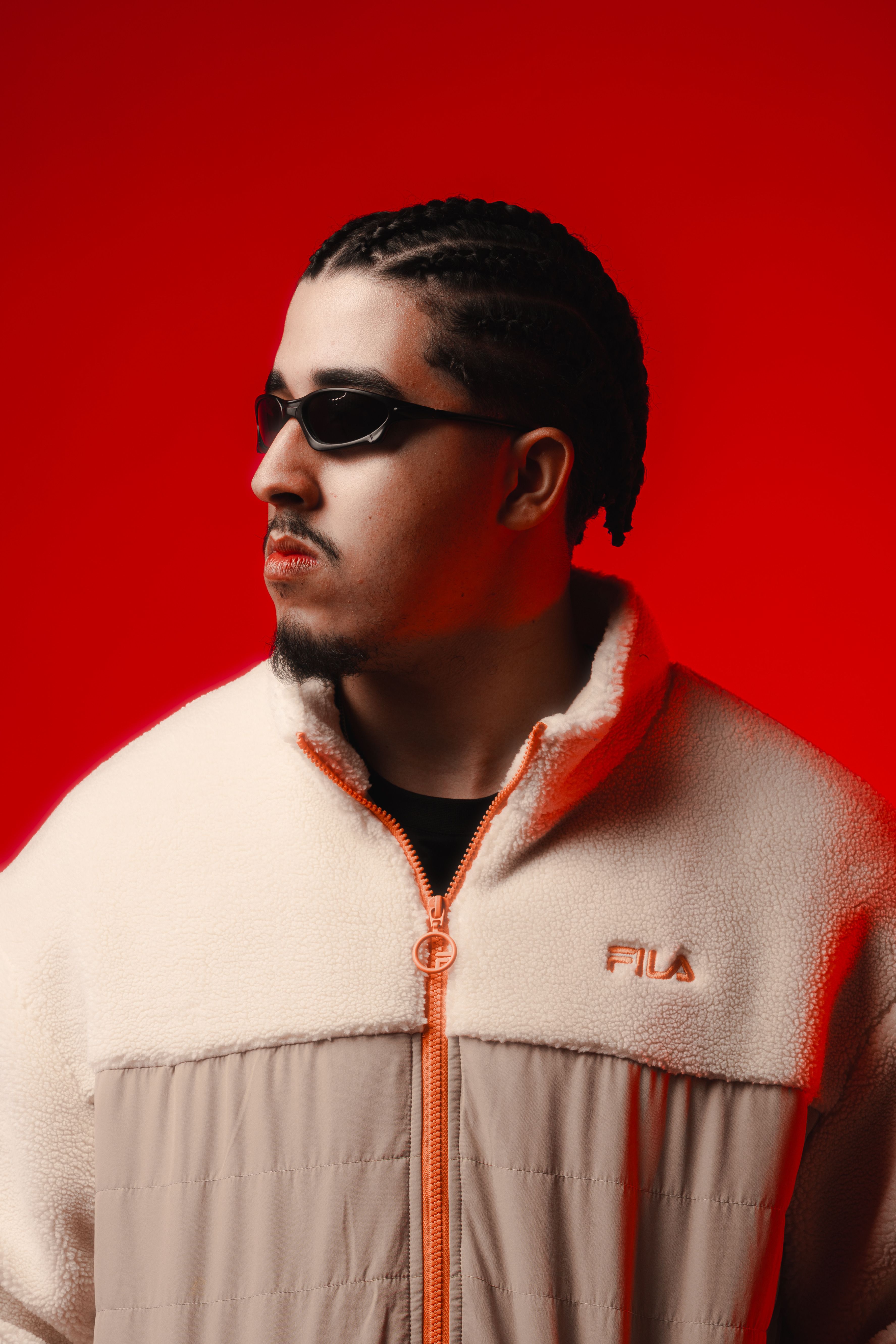 Portrait of a man with braided hair wearing sunglasses and a jacket, set against a vibrant red background.