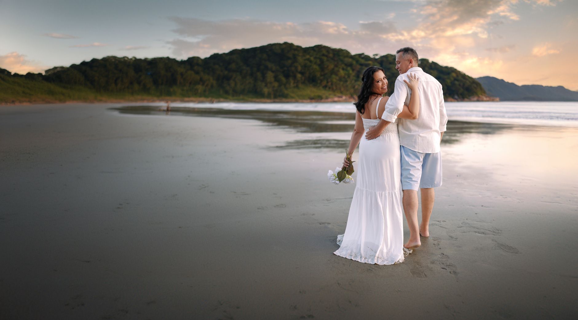 bride standing at the edge of a white-sand tropical beach, wearing effortless boho waves with three small fresh tropical blooms scattered through her hair, candid moment looking out at the ocean, gold
