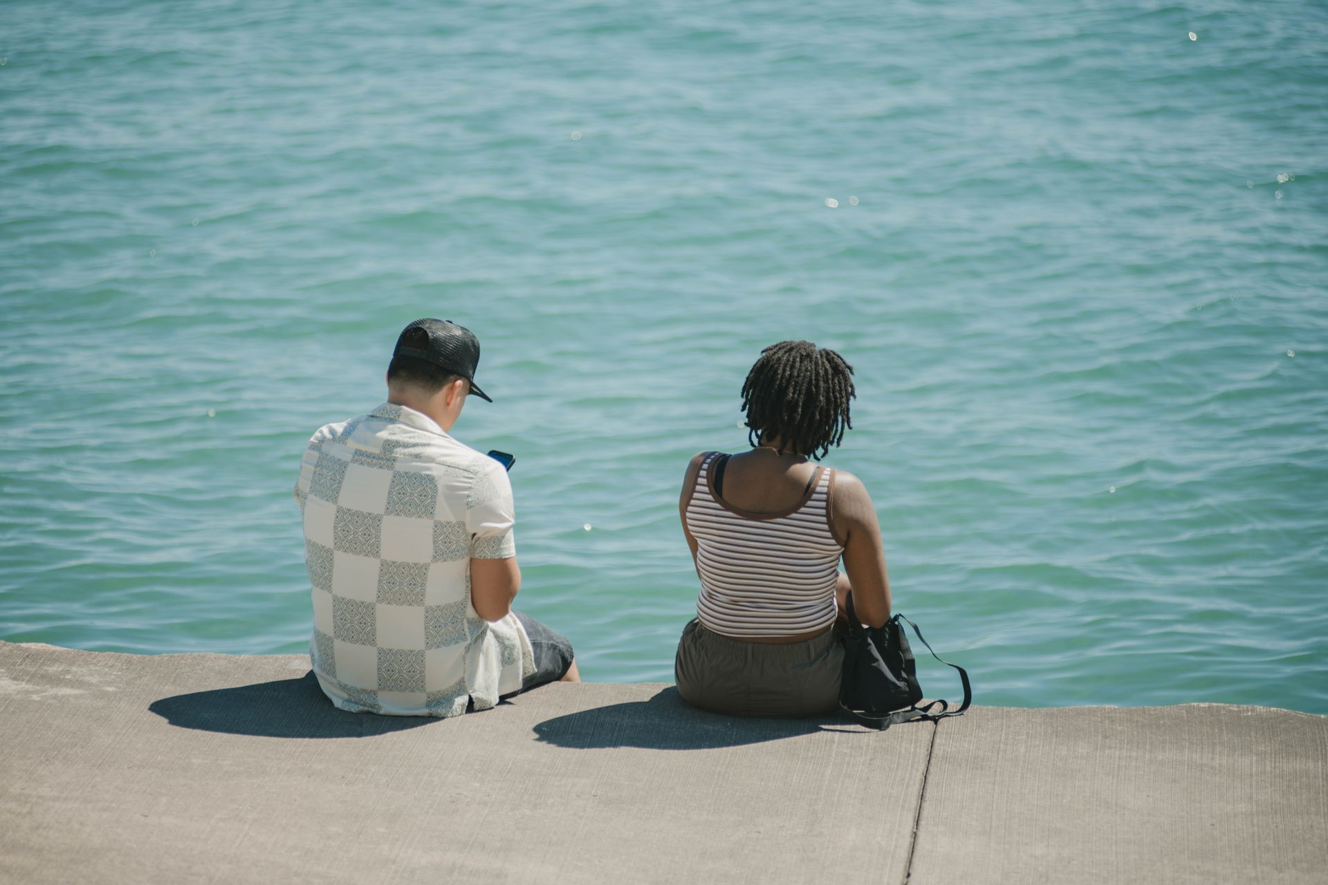 Two individuals sit on a concrete shore overlooking a serene lake in Chicago on a sunny day.