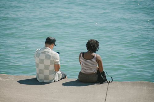 Free Two individuals sit on a concrete shore overlooking a serene lake in Chicago on a sunny day. Stock Photo